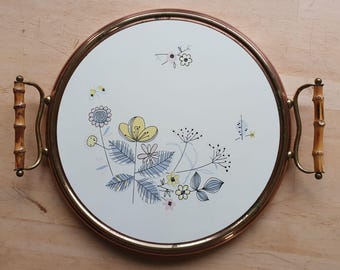 Op de afbeelding: Een ronde dienblad met een koperen rand en bamboe handvatten. Het dienblad heeft een witte achtergrond met een bloemenmotief in tinten blauw, geel en roze.