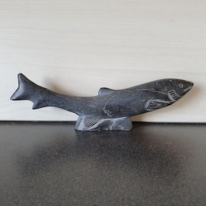 Op de afbeelding: Een donkergrijze stenen sculptuur van een vis. De vis is gebeeldhouwd met gedetailleerde vinnen en schubben en is gemonteerd op een kleine, gebeeldhouwde basis. De sculptuur wordt tentoongesteld op een donker oppervlak met een lichte houten achtergrond.