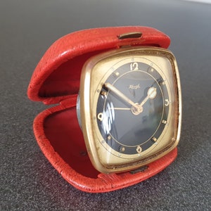 May include: A gold-colored alarm clock with a black face and white numerals. The clock is in a red leather case.
