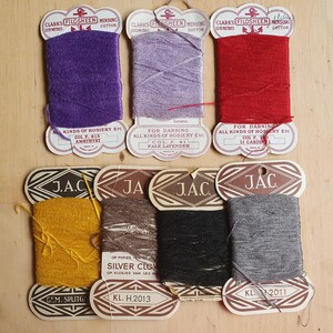 May include: Six spools of thread with labels for darning and mending. The spools are labelled with colours like amethyst, pale lavender, and cardinal. The thread is wound on cardboard spools with a J.A.C. label.