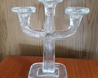 May include: Clear glass candelabra with three candle holders. The candelabra has a square base and a central stem that branches out to hold the candle cups. The design features a textured surface and a decorative floral element.