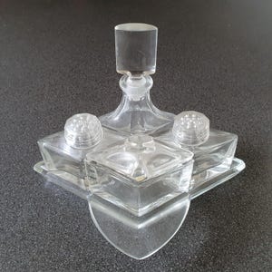 Può includere: Set da toeletta in vetro trasparente con una bottiglia di profumo centrale e due piccoli vasetti con coperchi. Il set ha una base a forma di cuore e un vassoio rettangolare. La bottiglia di profumo ha un tappo cilindrico.