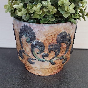 May include: A ceramic planter with a brown, white, and green glaze. The planter has a decorative design of black and green swirls. A small green plant is in the planter.