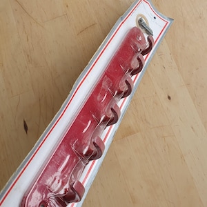 Może przedstawiać: Czerwony wieszak ścienny z pięcioma zakrzywionymi haczykami. Wieszak jest zapakowany w przezroczysty plastik i przymocowany do kartonowego podkładu za pomocą śruby. Haczyki są w ciemniejszym odcieniu czerwieni.