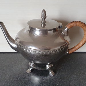 Op de afbeelding: Een zilveren theepot met een gevlochten handvat en een decoratief bladerpatroon rond de basis. De theepot heeft een deksel met een knop en staat op drie sierlijke voetjes. De handgreep is gemaakt van gevlochten bruin materiaal.