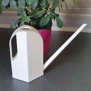 Pode incluir: Um regador branco, de formato geométrico, com um bico longo e fino. O regador tem uma grande abertura para a alça e fica sobre uma superfície escura. Um vaso rosa com uma planta verde está ao fundo.