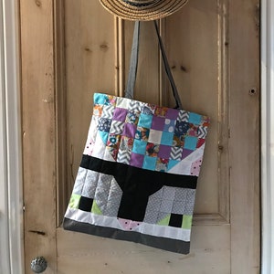 May include: A colorful patchwork tote bag with a black and white geometric design hangs on a wooden door with a straw hat above it.