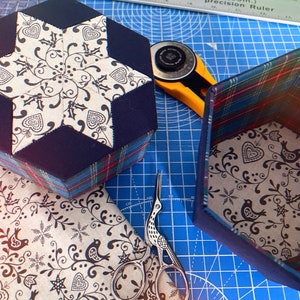 May include: Two hexagonal boxes with a star-shaped design on the top. The boxes are covered in a blue and red plaid fabric. The box on the left has a white star with a black floral pattern. The box on the right has a white floral pattern with black birds and hearts.