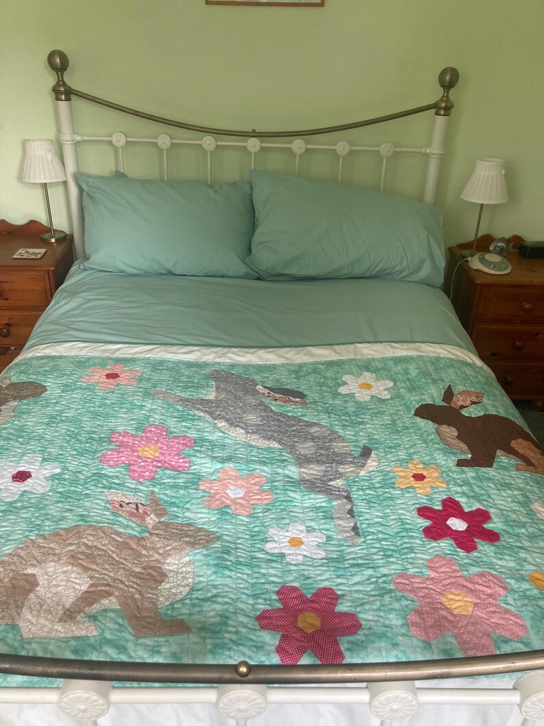 Op de afbeelding: Een turquoiseblauwe deken met een bloemenpatroon en twee bruine konijnen. De deken ligt op een wit metalen bedframe met een groene hoofdeinde.