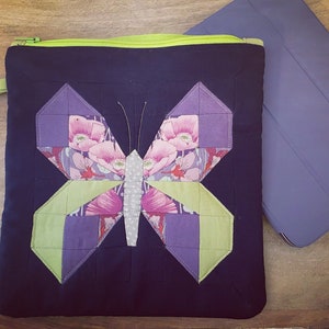 May include: A navy blue quilted fabric tablet case with a butterfly design. The butterfly has purple, green, and floral patterned wings. The case has a green zipper and a small green handle. A purple tablet case is next to it.