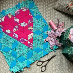 Puede incluir: Un bloque de colcha con un diseño de corazón rosa rodeado de un borde azul turquesa con un patrón floral. El bloque está rodeado de otras piezas de colcha, un carrete de hilo, tijeras y una estrella de tela verde y morada.