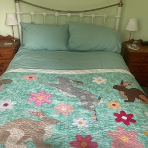Pode incluir: Um edredom azul turquesa com um padr&atilde;o floral e dois coelhos marrons. O edredom est&aacute; em uma estrutura de cama de metal branco com uma cabeceira verde.