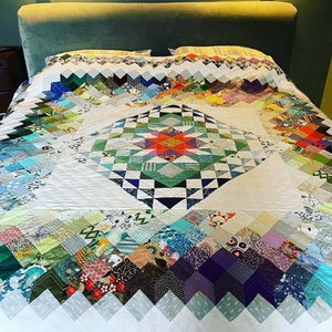 May include: A white quilt with a large star-shaped pattern in the centre. The quilt is made of various colourful fabrics, including green, blue, red, and yellow. The quilt is laid out on a bed with a green headboard.