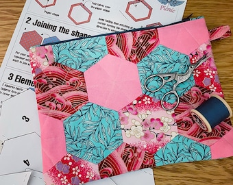 May include: A pink and blue quilted pouch with a zipper closure. The pouch is made of fabric with a floral pattern and is surrounded by paper templates numbered 1, 3, and 4. The pouch is lying on a wooden surface.