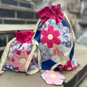 May include: Two drawstring bags and a coaster with a floral patchwork design. The bags have a pink top and drawstring, with a gold ribbon drawstring. The patchwork features pink, blue, and white floral patterns.