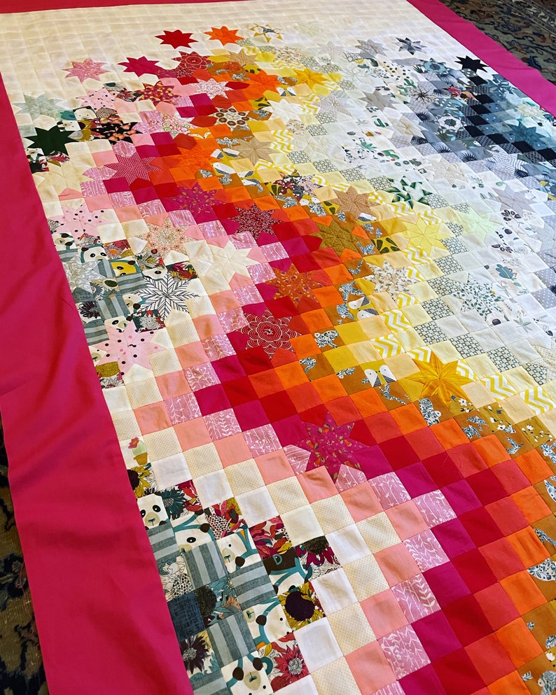 Op de afbeelding: Een kleurrijk quilt met een verloop van roze, oranje, geel en wit. De quilt is gemaakt van kleine vierkanten en sterren, en de randen zijn afgewerkt met een felroze stof.