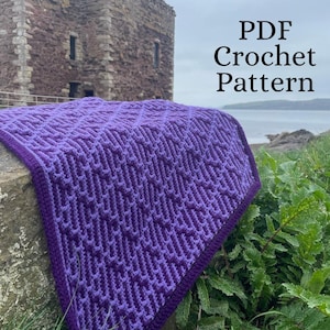 Może przedstawiać: Fioletowy koc szydełkowy z geometrycznym wzorem, narzucony na kamienną powierzchnię. Koc ma ciemnofioletową obwódkę. Tekst "PDF Crochet Pattern" jest widoczny w prawym górnym rogu. W tle kamienny budynek i morze.