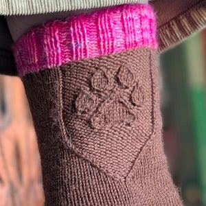 May include: Brown knitted sock with a pink cuff, heel, and toe. The sock has a textured paw print design on the ankle. The sock is worn on a foot, with a neutral-coloured carpet in the foreground.