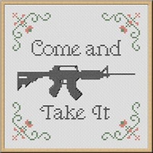 May include: Cross-stitch art with a wooden frame. The design features a black rifle silhouette and the text "Come and Take It" in black. Floral accents in green and pink adorn the corners, set against a white background.