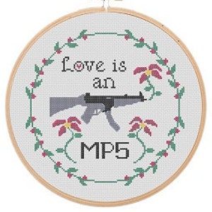 May include: Cross-stitch embroidery hoop with a white background and a floral border. The center of the hoop features a gray rifle with the text "Love is an MP5" in black.