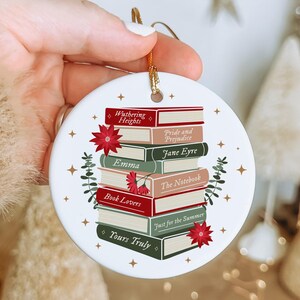 May include: A round white ceramic ornament featuring a stack of colourful books with titles including Wuthering Heights, Pride and Prejudice, Jane Eyre, Emma, The Notebook, Book Lovers, Just for the Summer, and Yours Truly, accented by red flowers.