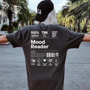 May include: A black t-shirt with white text that reads "100% TBR Reading Full" and "Mood Reader". The shirt also has a graphic of a barcode and a list of icons.
