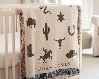 May include: A cream-coloured woven throw blanket featuring dark brown illustrations of a snake, star, horseshoe, bison, cactus, bull skull, cowboy hat, and a person riding a horse with a lasso. The name LUCAS JAMES is printed in dark brown text.
