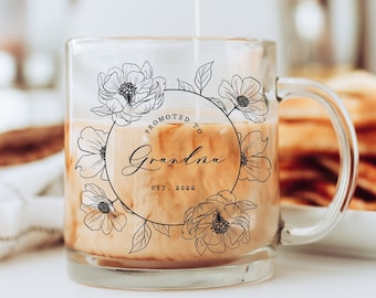 May include: A clear glass mug with a black floral design and the text "Promoted to Grandma Est. 2023" printed on it. The mug is filled with coffee and milk.