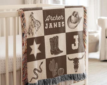 May include: A woven throw blanket featuring a grid pattern with brown and cream squares. Illustrations include a cowboy on a bucking horse, a cowboy hat, a sheriff star, cowboy boots, a letter J, a snake, cacti, and a bull skull. The text reads Archer James.