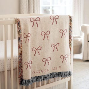 May include: A cream-colored woven throw blanket featuring a repeating pattern of dark pink bows and the name OLIVIA LILY printed in dark pink text. The blanket has a fringed edge with multicolored threads along the vertical sides.