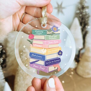 May include: A clear, circular Christmas decoration depicting a pile of books with floral decorations. The book titles include "Wuthering Heights," "Pride and Prejudice," and "Emma." It has a gold ribbon for hanging and the words "ink + luxe" at the bottom.