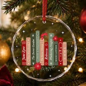 May include: A clear, circular, faceted glass ornament hanging from a red ribbon. The centre features a graphic of colourful book spines titled Jane Eyre, The Notebook, The Secret Garden, Pride & Prejudice, The Secret History, Rebecca, Wuthering Heights, Book Lovers, 1984, and Emma, accented with red poinsettia flowers and small white stars.