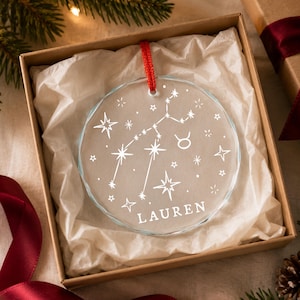 May include: A circular clear glass ornament featuring a white etched Taurus zodiac constellation, the Taurus symbol, and the name LAUREN. The ornament hangs from a red ribbon and is presented inside a square cardboard gift box with white tissue paper.