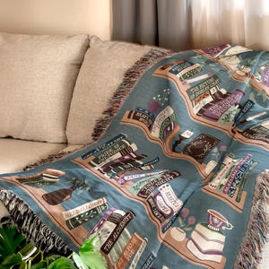 May include: A teal throw blanket featuring a bookshelf design with classic literature titles like "Pride & Prejudice" and "The Picture of Dorian Gray." The blanket has a fringed edge and is draped over a beige sofa.