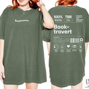 K&ouml;nnte beinhalten: Ein gr&uuml;nes T-Shirt mit dem Aufdruck "Booktrovert" auf der Vorderseite. Die R&uuml;ckseite des Shirts zeigt eine Grafik mit dem Text "100% TBR Reading Full Book-trovert" und eine Liste der Zutaten f&uuml;r einen Booktrovert.