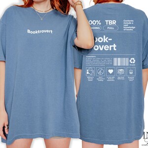 K&ouml;nnte beinhalten: Ein blaues T-Shirt mit dem Text "Booktrovert" auf der Vorderseite und "100% TBR, Reading Full, Book-overt" auf der R&uuml;ckseite. Die R&uuml;ckseite enth&auml;lt auch eine Liste von Zutaten, um ein Booktrovert zu sein, darunter "bereit, B&uuml;cher zu entdecken", "erwarte kein schnelles Lesen", "verliebt in fiktive Charaktere", "habe &uuml;ber 1000 B&uuml;cher geliebt" und "ben&ouml;tigt Lesesaft".