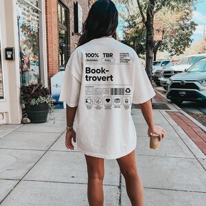 K&ouml;nnte beinhalten: Ein wei&szlig;es T-Shirt mit schwarzem Text, der "100% TBR Lesen Vollst&auml;ndig Book-trovert" lautet und eine Liste von Merkmalen eines Book-troverts enth&auml;lt. Der Text sagt auch "Inhalt umfasst ein umfassendes Wissen &uuml;ber B&uuml;cher."