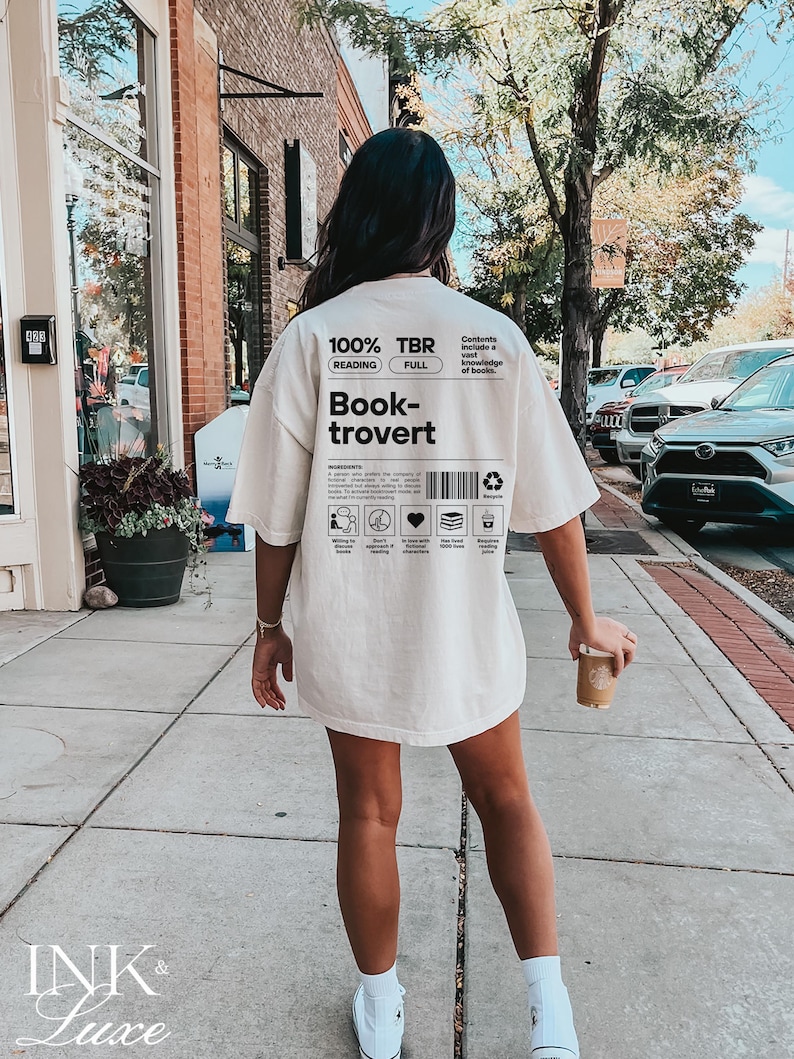 K&ouml;nnte beinhalten: Ein wei&szlig;es T-Shirt mit schwarzem Text, der "100% TBR Lesen Vollst&auml;ndig Book-trovert" lautet und eine Liste von Merkmalen eines Book-troverts enth&auml;lt. Der Text sagt auch "Inhalt umfasst ein umfassendes Wissen &uuml;ber B&uuml;cher."