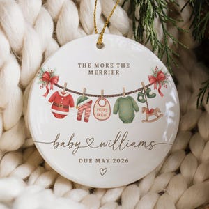 May include: A round white ceramic ornament featuring a clothesline illustration with baby clothes, a bib, and a rocking horse. Text reads THE MORE THE MERRIER, baby Williams, and DUE MAY 2026. Red bows and greenery accents decorate the top corners.