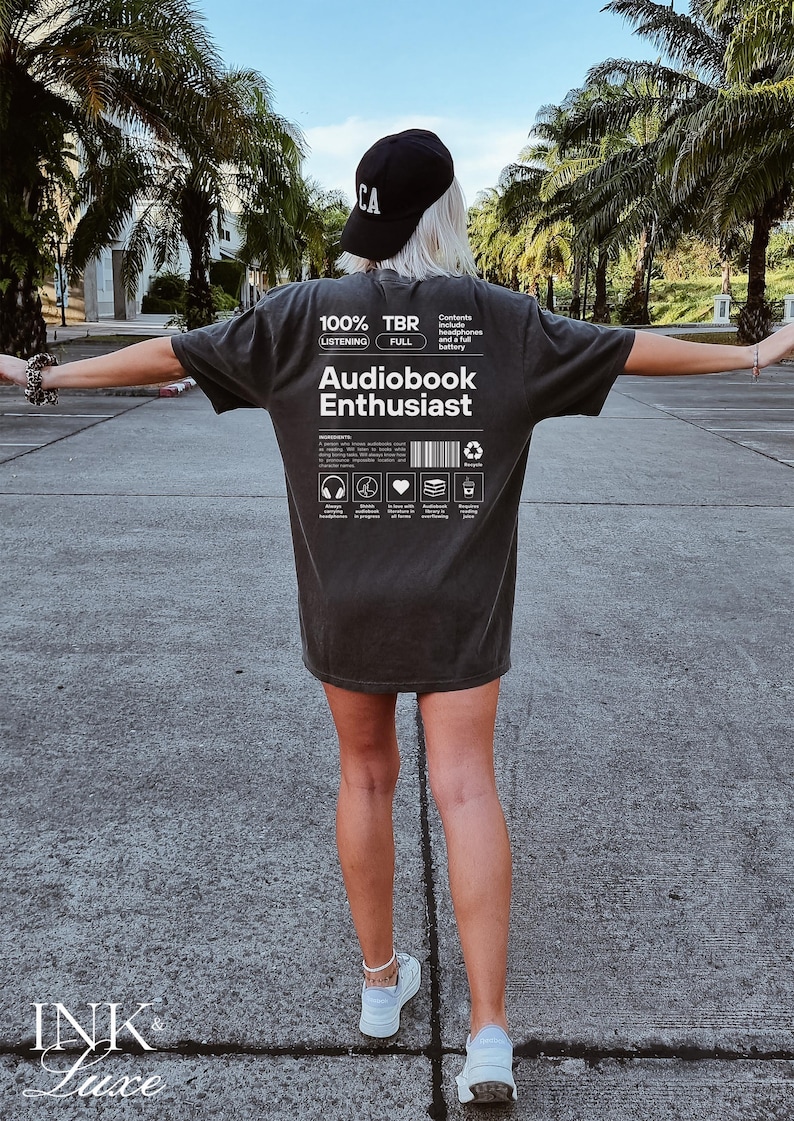 May include: A person wearing a dark grey t-shirt with the text "100% Listening TBR Full Audiobook Enthusiast" printed on the back. The text includes icons for headphones, a heart, a stack of books, and a recycling symbol.
