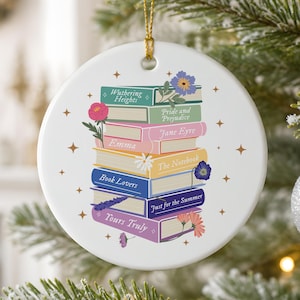 May include: A round white ceramic ornament featuring a stack of colorful books with titles including Wuthering Heights, Pride and Prejudice, Jane Eyre, Emma, The Notebook, Book Lovers, Just for the Summer, and Yours Truly, decorated with floral accents.