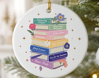 May include: A round white ceramic ornament featuring a stack of colorful books with titles including Wuthering Heights, Pride and Prejudice, Jane Eyre, Emma, The Notebook, Book Lovers, Just for the Summer, and Yours Truly, decorated with floral accents.