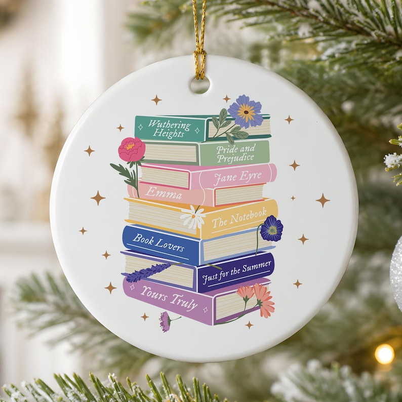 May include: A round white ceramic ornament featuring a stack of colorful books with titles including Wuthering Heights, Pride and Prejudice, Jane Eyre, Emma, The Notebook, Book Lovers, Just for the Summer, and Yours Truly, decorated with floral accents.