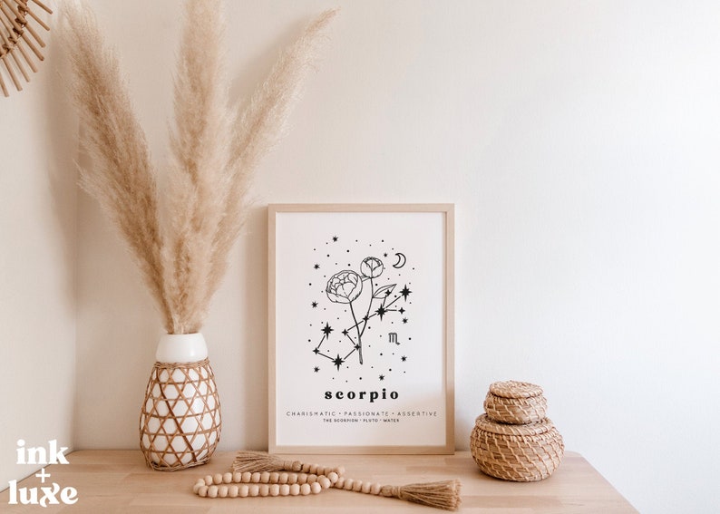 May include: A framed art print featuring a black and white floral design with the word "Scorpio" and related text. The print is displayed on a wooden surface with decorative items, including a vase with pampas grass and woven storage baskets.