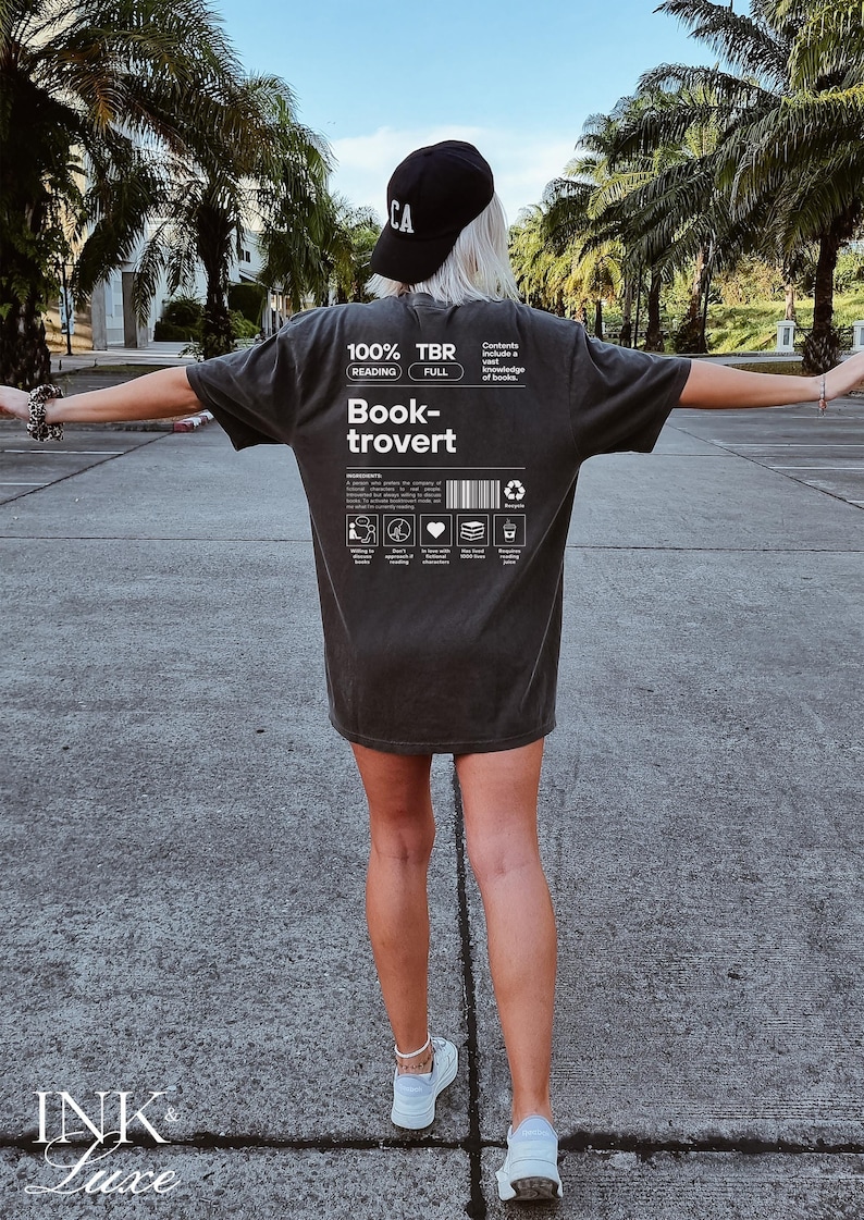 K&ouml;nnte beinhalten: Ein dunkelgraues T-Shirt mit wei&szlig;em Text, der "100% TBR Lesen Vollst&auml;ndig Book-trovert" lautet und einer Grafik eines Buches mit einem Herzen, einem Stapel B&uuml;chern und einem Recycling-Symbol. Der Text "Inhalt Umfasst ein umfassendes Wissen &uuml;ber B&uuml;cher." ist ebenfalls auf dem T-Shirt gedruckt.