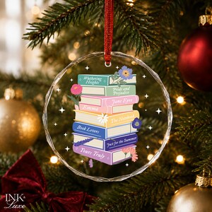 May include: A circular, faceted glass ornament hanging from a red ribbon, featuring a colorful illustration of a stack of books with titles including Wuthering Heights, Pride and Prejudice, Jane Eyre, Emma, The Notebook, Book Lovers, Just for the Summer, and Yours Truly, accented with small floral drawings and white star icons.