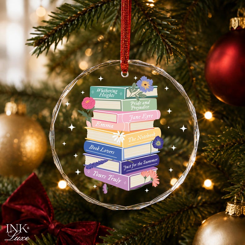 May include: A circular, faceted glass ornament hanging from a red ribbon, featuring a colorful illustration of a stack of books with titles including Wuthering Heights, Pride and Prejudice, Jane Eyre, Emma, The Notebook, Book Lovers, Just for the Summer, and Yours Truly, accented with small floral drawings and white star icons.