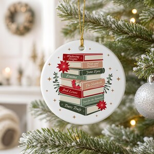 May include: A round white ceramic ornament featuring a stack of books with titles including Wuthering Heights, Pride and Prejudice, Jane Eyre, Emma, The Notebook, Book Lovers, Just for the Summer, and Yours Truly, accented with red poinsettias and gold stars.