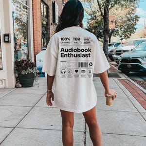 May include: A white t-shirt with black text that reads "100% TBR Listening Full" and "Audiobook Enthusiast". The text is in a box with a barcode and other symbols.