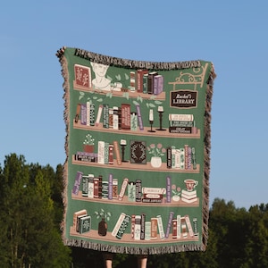 May include: A green woven throw blanket with a bookshelf design featuring a variety of book titles. The blanket has a fringe border and the text "Rachel's Library" is displayed on a sign hanging from the top shelf.